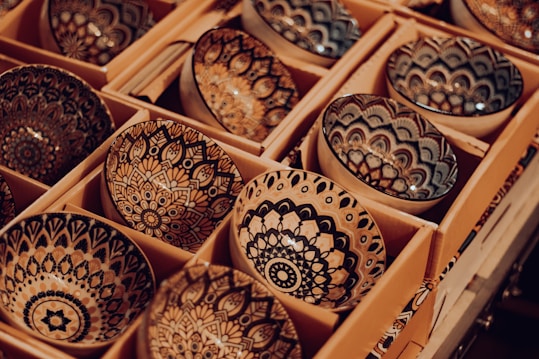 A collection of intricately designed ceramic bowls arranged in rows within cardboard packaging. Each bowl features detailed, symmetrical patterns in shades of black, tan, and white, creating an artistic and handcrafted appearance.