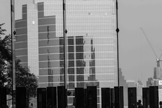 A black and white image featuring a large glass-panel skyscraper surrounded by other high-rise buildings. Shadows of people are visible in the lower part of the image, along with a tree on the left side. A construction crane is visible to the right, adding to the urban landscape.