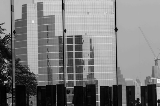 A black and white image featuring a large glass-panel skyscraper surrounded by other high-rise buildings. Shadows of people are visible in the lower part of the image, along with a tree on the left side. A construction crane is visible to the right, adding to the urban landscape.