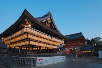 A traditional Japanese shrine adorned with rows of lanterns, featuring intricate architectural details and illuminated softly against the backdrop of an early evening sky. A few people are walking around the shrine area, giving a sense of timeless tranquility.
