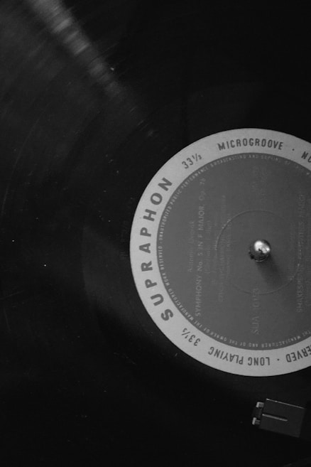 A close-up of a vinyl record on a turntable, featuring visible grooves and a label with the text 'SUPRAPHON' and 'MICROGROOVE'. The image is in black and white, focusing on the circular label and the tonearm.