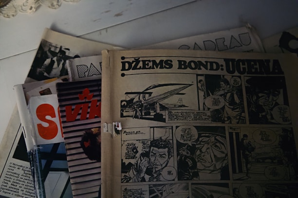 A collection of vintage comic books is spread out on a surface. The comic on top features the title 'DŽEMS BOND: UCENA' with illustrations and speech bubbles in a comic strip style. Other partially visible publications beneath include text and graphics, with one showing part of a word 'VIK'. The overall setting suggests a retro or nostalgic theme.