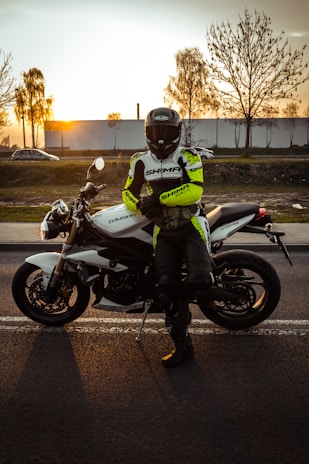 A motorcyclist wearing a helmet and protective gear stands confidently beside a white motorcycle on the road. The background features a sunset, casting warm hues over the scene, with silhouettes of bare trees and distant buildings visible against the evening sky.