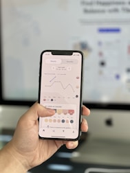 A person holds a smartphone displaying a graph and color-coded schedule on the screen. The background is slightly blurred, showing a computer monitor.