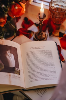 An open book with text and an image of a person's back, surrounded by red rose petals, an ornate glass with liquid, and a lit candle in a brass holder on a table. Bright orange flowers are partially visible at the edges, creating a warm and cozy atmosphere.