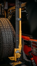 A mechanical jack with a yellow handle is mounted next to a spare tire on the rear of a red vehicle. The jack has multiple adjustment holes along its vertical frame. A bottle of wheel cleaner is visible next to it.