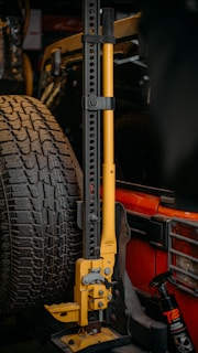 A mechanical jack with a yellow handle is mounted next to a spare tire on the rear of a red vehicle. The jack has multiple adjustment holes along its vertical frame. A bottle of wheel cleaner is visible next to it.