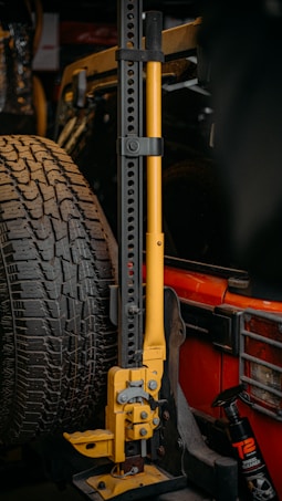 A mechanical jack with a yellow handle is mounted next to a spare tire on the rear of a red vehicle. The jack has multiple adjustment holes along its vertical frame. A bottle of wheel cleaner is visible next to it.