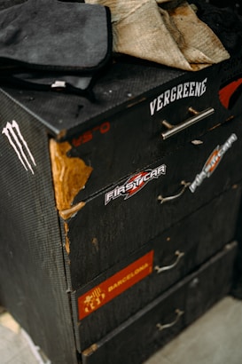 A worn, black dresser with several drawers, showing signs of wear such as scratches and peeling paint. It is decorated with various stickers, including those for 'Firstcar' and 'Barcelona'. Items like cloths and a black case are placed on top of the dresser.