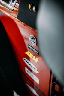 A close-up view of a red vehicle panel adorned with multiple brand stickers, including AEM and Trail Rated 4x4. The focus is on the stickers with some parts of the vehicle's interior visible.