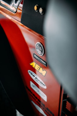 A close-up view of a red vehicle panel adorned with multiple brand stickers, including AEM and Trail Rated 4x4. The focus is on the stickers with some parts of the vehicle's interior visible.