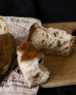 A close-up shot of partially eaten artisan bread placed on a piece of newspaper and a wooden cutting board with a dark background. The bread has a golden brown crust and an airy, porous interior.
