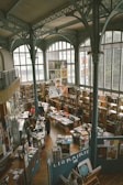A spacious bookstore with high ceilings and large arched windows, filled with bookshelves and tables displaying various books and printed materials. People are browsing and engaging with the books. The architectural design includes ornate metal beams and columns. Posters and artwork are displayed throughout.