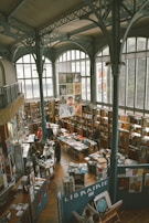A spacious bookstore with high ceilings and large arched windows, filled with bookshelves and tables displaying various books and printed materials. People are browsing and engaging with the books. The architectural design includes ornate metal beams and columns. Posters and artwork are displayed throughout.