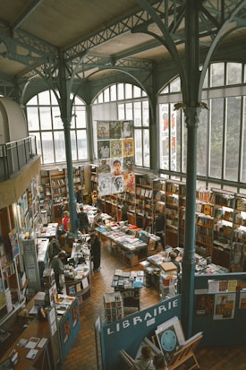A spacious bookstore with high ceilings and large arched windows, filled with bookshelves and tables displaying various books and printed materials. People are browsing and engaging with the books. The architectural design includes ornate metal beams and columns. Posters and artwork are displayed throughout.