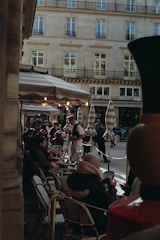 A bustling outdoor cafe with people sitting at tables under striped umbrellas. A waiter is serving drinks, and the surrounding architecture features a classic European facade with multiple windows.