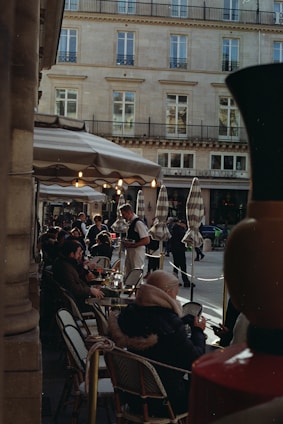 A bustling outdoor cafe with people sitting at tables under striped umbrellas. A waiter is serving drinks, and the surrounding architecture features a classic European facade with multiple windows.