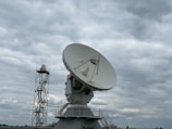 A large satellite dish is prominently displayed against a cloudy sky. The dish is mounted on a sturdy structure with a staircase leading up to it, suggesting it is part of a communications or research facility. Beside it, a tall metal tower is visible.