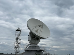A large satellite dish is prominently displayed against a cloudy sky. The dish is mounted on a sturdy structure with a staircase leading up to it, suggesting it is part of a communications or research facility. Beside it, a tall metal tower is visible.