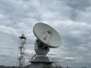 A large satellite dish is prominently displayed against a cloudy sky. The dish is mounted on a sturdy structure with a staircase leading up to it, suggesting it is part of a communications or research facility. Beside it, a tall metal tower is visible.