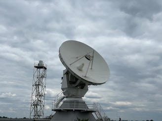 A large satellite dish is prominently displayed against a cloudy sky. The dish is mounted on a sturdy structure with a staircase leading up to it, suggesting it is part of a communications or research facility. Beside it, a tall metal tower is visible.