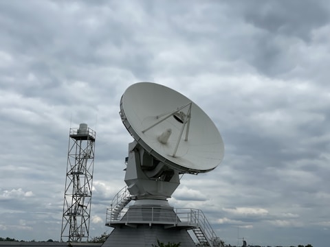 A large satellite dish is prominently displayed against a cloudy sky. The dish is mounted on a sturdy structure with a staircase leading up to it, suggesting it is part of a communications or research facility. Beside it, a tall metal tower is visible.