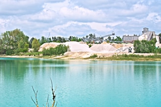 A serene landscape featuring a body of turquoise water in the foreground with a shoreline covered in greenery. Beyond the water, there are piles of sand and an industrial setup with several structures and machinery. The sky above is partly cloudy, adding a calm atmosphere to the scene.
