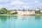 A serene landscape featuring a body of turquoise water in the foreground with a shoreline covered in greenery. Beyond the water, there are piles of sand and an industrial setup with several structures and machinery. The sky above is partly cloudy, adding a calm atmosphere to the scene.