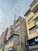 Tall, weathered buildings with prominent signs line a street that is covered by a web of overhead cables against a cloudy sky. The structures have signs like Hotel Al Haram and brands like Sony, indicating a mix of residential and commercial use.
