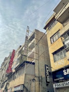 Tall, weathered buildings with prominent signs line a street that is covered by a web of overhead cables against a cloudy sky. The structures have signs like Hotel Al Haram and brands like Sony, indicating a mix of residential and commercial use.