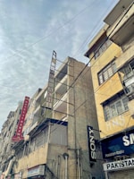 Tall, weathered buildings with prominent signs line a street that is covered by a web of overhead cables against a cloudy sky. The structures have signs like Hotel Al Haram and brands like Sony, indicating a mix of residential and commercial use.