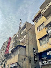 Tall, weathered buildings with prominent signs line a street that is covered by a web of overhead cables against a cloudy sky. The structures have signs like Hotel Al Haram and brands like Sony, indicating a mix of residential and commercial use.