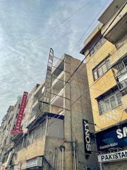 Tall, weathered buildings with prominent signs line a street that is covered by a web of overhead cables against a cloudy sky. The structures have signs like Hotel Al Haram and brands like Sony, indicating a mix of residential and commercial use.