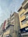 Tall, weathered buildings with prominent signs line a street that is covered by a web of overhead cables against a cloudy sky. The structures have signs like Hotel Al Haram and brands like Sony, indicating a mix of residential and commercial use.