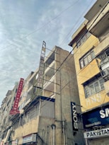 Tall, weathered buildings with prominent signs line a street that is covered by a web of overhead cables against a cloudy sky. The structures have signs like Hotel Al Haram and brands like Sony, indicating a mix of residential and commercial use.