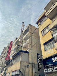 Tall, weathered buildings with prominent signs line a street that is covered by a web of overhead cables against a cloudy sky. The structures have signs like Hotel Al Haram and brands like Sony, indicating a mix of residential and commercial use.