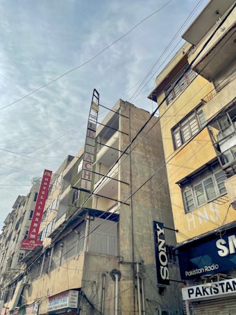 Tall, weathered buildings with prominent signs line a street that is covered by a web of overhead cables against a cloudy sky. The structures have signs like Hotel Al Haram and brands like Sony, indicating a mix of residential and commercial use.