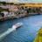 A large white cruise ship travels along a wide river with a wake trailing behind it. The river is flanked by steep rocky cliffs topped with colorful buildings and greenery. A prominent yellow bridge spans across the river in the background, and the scene is bathed in warm sunlight.