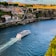 A large white cruise ship travels along a wide river with a wake trailing behind it. The river is flanked by steep rocky cliffs topped with colorful buildings and greenery. A prominent yellow bridge spans across the river in the background, and the scene is bathed in warm sunlight.