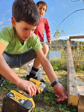 Two young boys are outdoors engaged in a woodworking activity. One boy is using a power drill on a piece of wood, while the other observes. The setting appears to be a backyard with grass and some scattered plants. Both are focused on their task, amidst the natural surroundings.