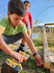 Two young boys are outdoors engaged in a woodworking activity. One boy is using a power drill on a piece of wood, while the other observes. The setting appears to be a backyard with grass and some scattered plants. Both are focused on their task, amidst the natural surroundings.