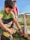 Two young boys are outdoors engaged in a woodworking activity. One boy is using a power drill on a piece of wood, while the other observes. The setting appears to be a backyard with grass and some scattered plants. Both are focused on their task, amidst the natural surroundings.
