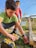 Two young boys are outdoors engaged in a woodworking activity. One boy is using a power drill on a piece of wood, while the other observes. The setting appears to be a backyard with grass and some scattered plants. Both are focused on their task, amidst the natural surroundings.