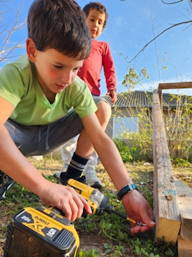 Two young boys are outdoors engaged in a woodworking activity. One boy is using a power drill on a piece of wood, while the other observes. The setting appears to be a backyard with grass and some scattered plants. Both are focused on their task, amidst the natural surroundings.