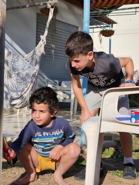 Two boys are outdoors near a hammock and a white plastic chair in a sunny yard. One boy crouches down while holding a red-handled paintbrush, focused on his task. The other boy stands nearby, watching with interest. The background features potted plants and a partially shaded patio.