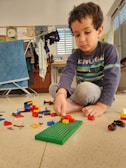A child is sitting on the floor playing with colorful building blocks. The child appears focused on assembling blocks on a green baseplate. Behind the child, there's a clothes rack with various garments and art displayed on a wall. Sunlight enters through a window with blinds, casting a warm glow on the scene.