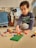 A child is sitting on the floor playing with colorful building blocks. The child appears focused on assembling blocks on a green baseplate. Behind the child, there's a clothes rack with various garments and art displayed on a wall. Sunlight enters through a window with blinds, casting a warm glow on the scene.