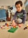 A child is sitting on the floor playing with colorful building blocks. The child appears focused on assembling blocks on a green baseplate. Behind the child, there's a clothes rack with various garments and art displayed on a wall. Sunlight enters through a window with blinds, casting a warm glow on the scene.