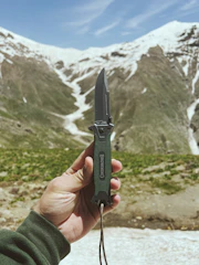 A sleek precision folding knife clipped to a hiker’s backpack strap in a mountain setting.