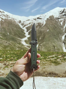 A sleek precision folding knife clipped to a hiker’s backpack strap in a mountain setting.
